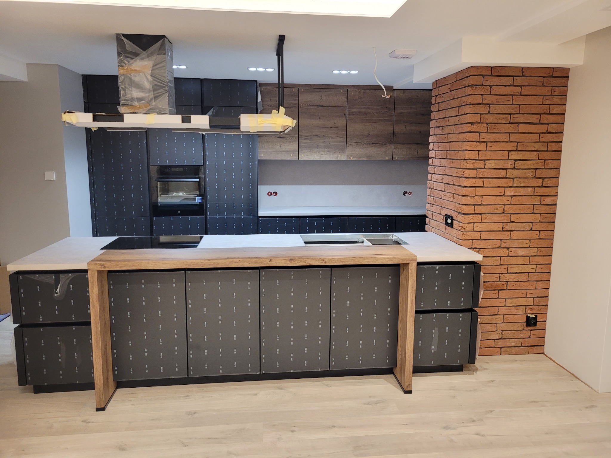 Nowoczesna kuchnia z wyspą i ceglaną ścianą, fronty szafek w folii ochronnej, okap częściowo zabezpieczony na czas montażu.