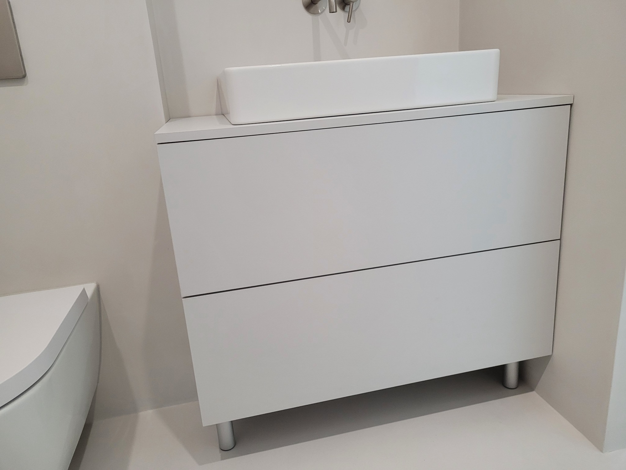 Minimalistyczna, biała szafka łazienkowa z dwiema szufladami i prostokątną umywalką nablatową, całość w jasnej, monochromatycznej kolorystyce.
