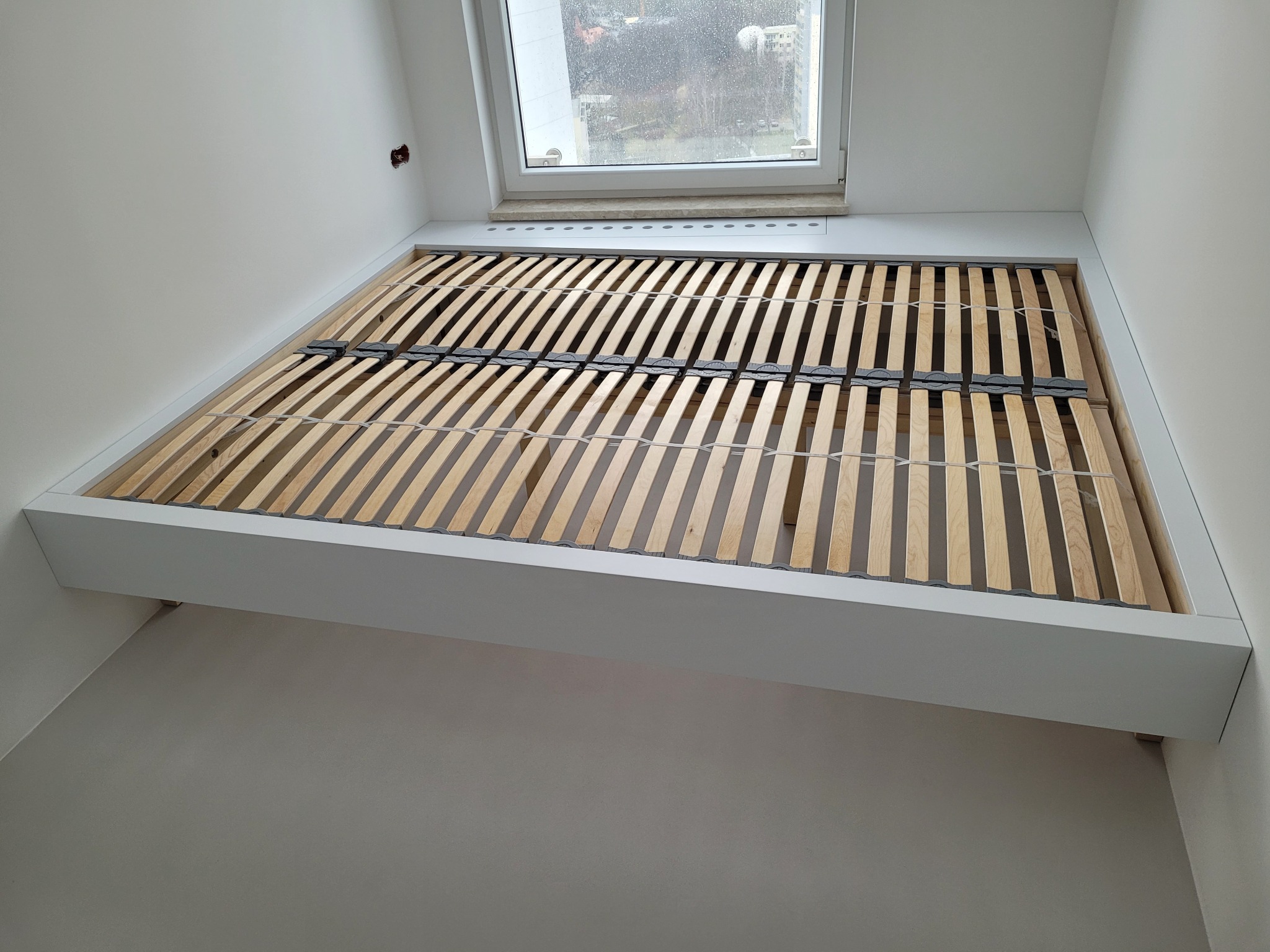 Rama łóżka z drewnianym stelażem, wbudowana w białą wnękę pod oknem, widok z dołu.