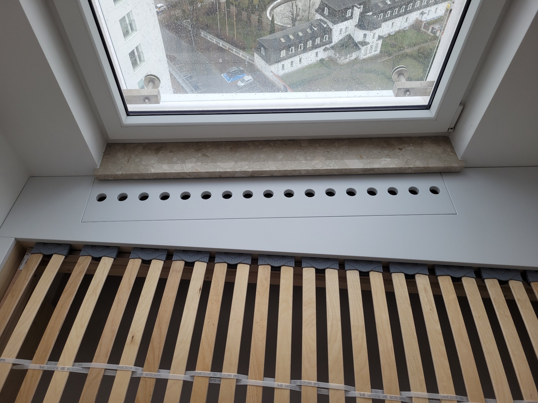 Okno dachowe z widokiem na deszczowy krajobraz miejski, kratka wentylacyjna poniżej, rama łóżka z drewnianymi listwami w dolnej części kadru.
