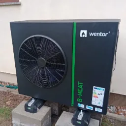 Czarna pompa ciepła Wentor B-HEAT ustawiona na betonowych bloczkach z regulowanymi wspornikami, z widoczną etykietą energetyczną i zielonym akcentem kolorystycznym.