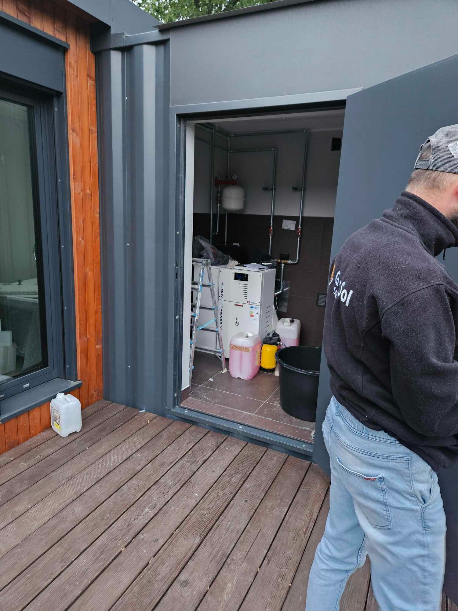 Instalacja pompy ciepła w pomieszczeniu technicznym z widocznymi rurami, zbiornikiem wyrównawczym i drabiną, pracownik w bluzie z logo firmy na tle wejścia do budynku z drewnianym tarasem.
