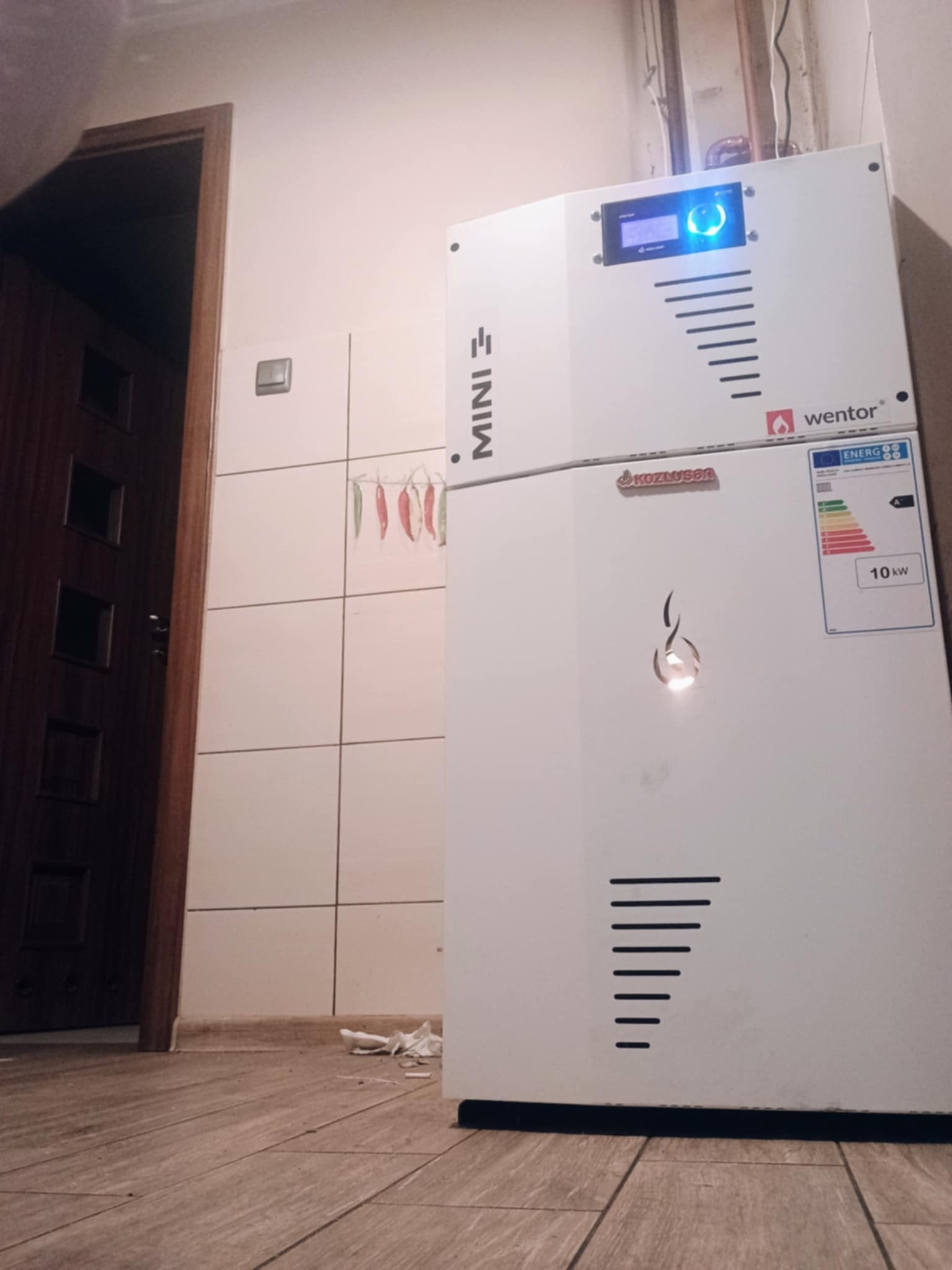 Biały, dwuczęściowy piec z wyświetlaczem i logotypami producentów Wentor i Kozlusan, zainstalowany w pomieszczeniu z brązowymi panelami podłogowymi, białymi kafelkami i suszonymi papryczkami chili...