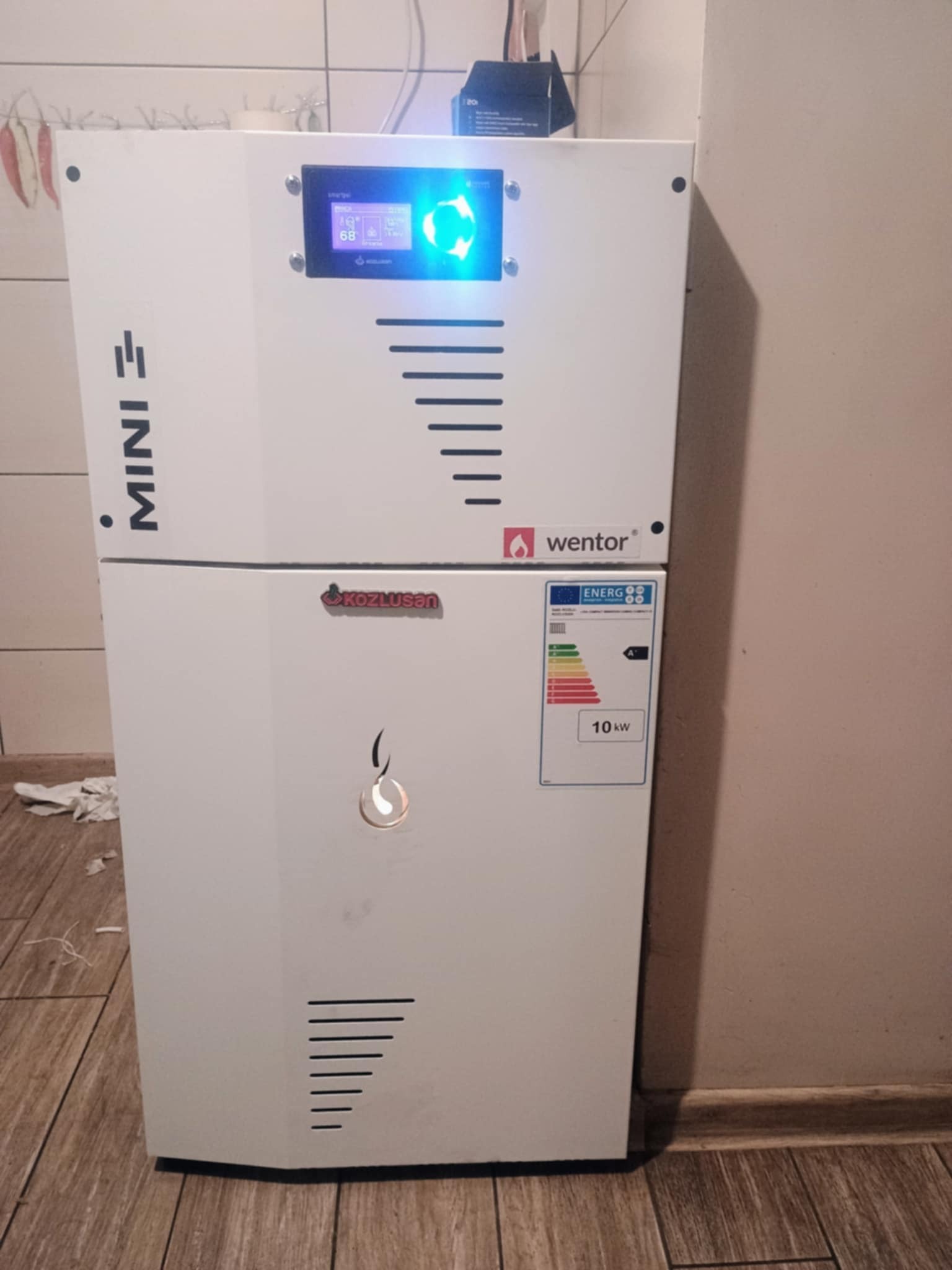 Biały, dwuczęściowy piec z wyświetlaczem i logotypami producentów Wentor i Kozlusan, stojący na brązowej podłodze z imitacją drewna, na tle beżowej ściany. Na górnej części pieca widoczny...