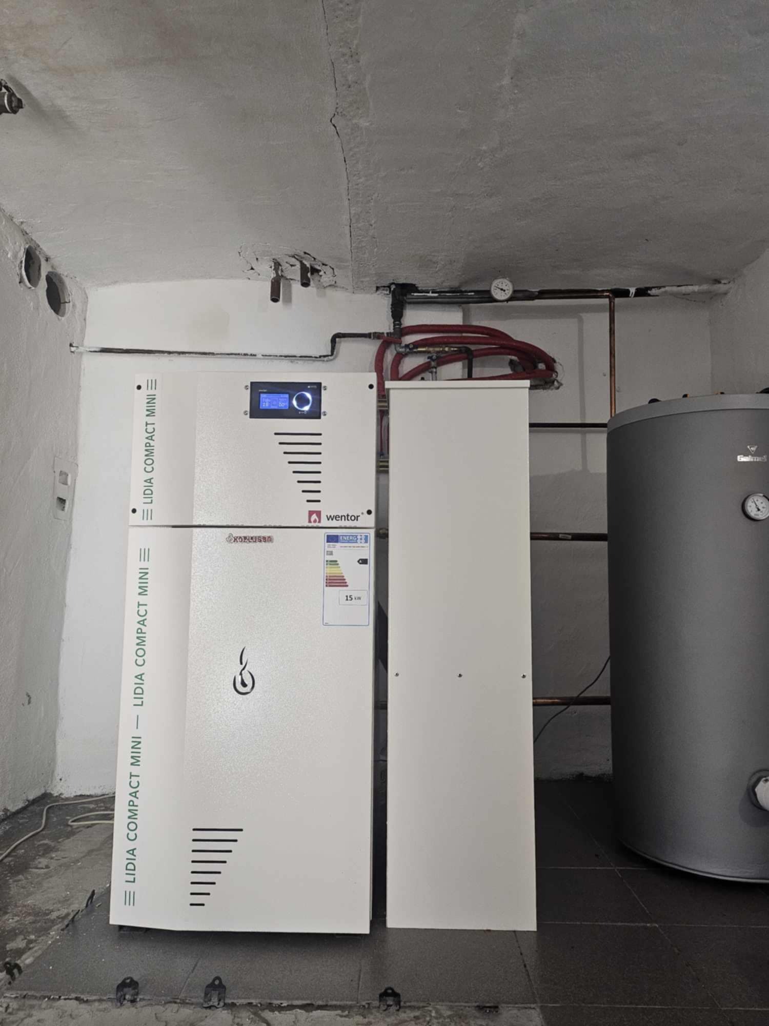 Kompaktowy, biały piec z wyświetlaczem i oznaczeniami producenta Wentor, zainstalowany w pomieszczeniu z szarą posadzką i widocznymi rurami instalacji grzewczej oraz zasobnikiem ciepłej wody.