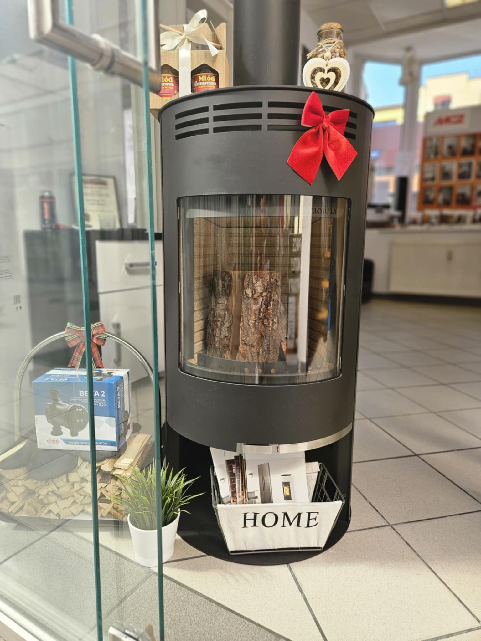 Czarny piec wolnostojący z szybą, udekorowany czerwoną kokardą i drewnianym sercem, prezentowany w salonie sprzedaży, obok gabloty z pompami obiegowymi i drewnem opałowym.