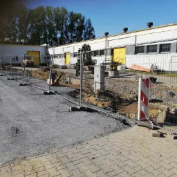 Teren budowy z wykopem zabezpieczonym siatką, widoczny sprzęt budowlany w tle, obok budynku przemysłowego z żółtymi elementami.