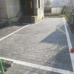 Ułożona szara kostka brukowa z białymi obrzeżami przed budynkiem, na nawierzchni leżą czerwone rękawice robocze.