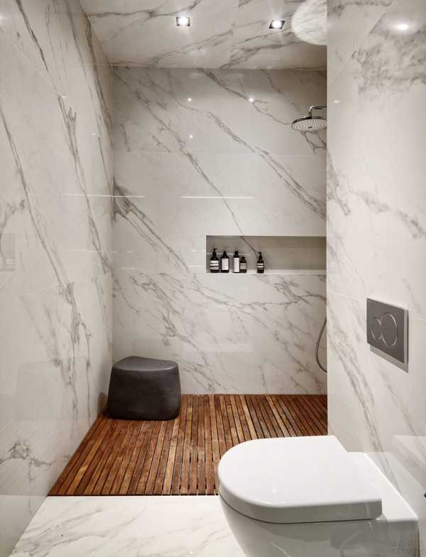 Nowoczesna łazienka z marmurowymi płytkami na ścianach, drewnianą podłogą w strefie prysznica i minimalistycznym wyposażeniem, w tym czarne butelki z kosmetykami w ściennej wnęce.