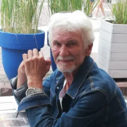 Uśmiechnięty mężczyzna z siwymi włosami w dżinsowej kurtce siedzi przy białym stoliku na zewnątrz, z donicami z trawą w tle.