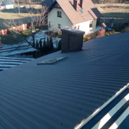 Montaż blachy trapezowej na dachu z oknem dachowym i kominem; widoczne elementy konstrukcji więźby dachowej.