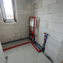 Surowy stan łazienki z widoczną instalacją podtynkową WC na czerwonym stelażu, rurami wodnymi z wyprowadzeniami na ścianie z betonu komórkowego oraz oknem z naklejkami producenta.