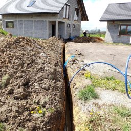 Mk Instal Plus - Wykop pod instalację, widoczny rów w ziemi z ułożonym niebieskim przewodem, w tle dwa budynki w stanie surowym, pracownik kopiący ziemię i rozrzucona ziemia.