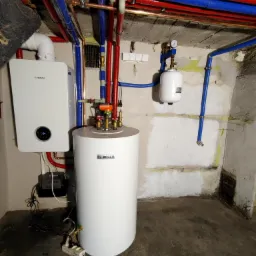 Nowoczesny, biały bojler i piec gazowy marki Bosch zamontowane na tle surowej ściany w piwnicy, widoczne rury w izolacji czerwonej i niebieskiej oraz manometr na ścianie.