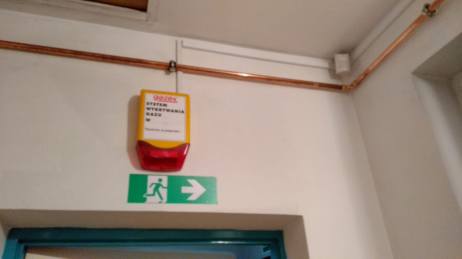 Miedziana instalacja gazowa pod sufitem, czujnik Gazex systemu wykrywania gazu oraz zielony znak ewakuacyjny na białej ścianie.