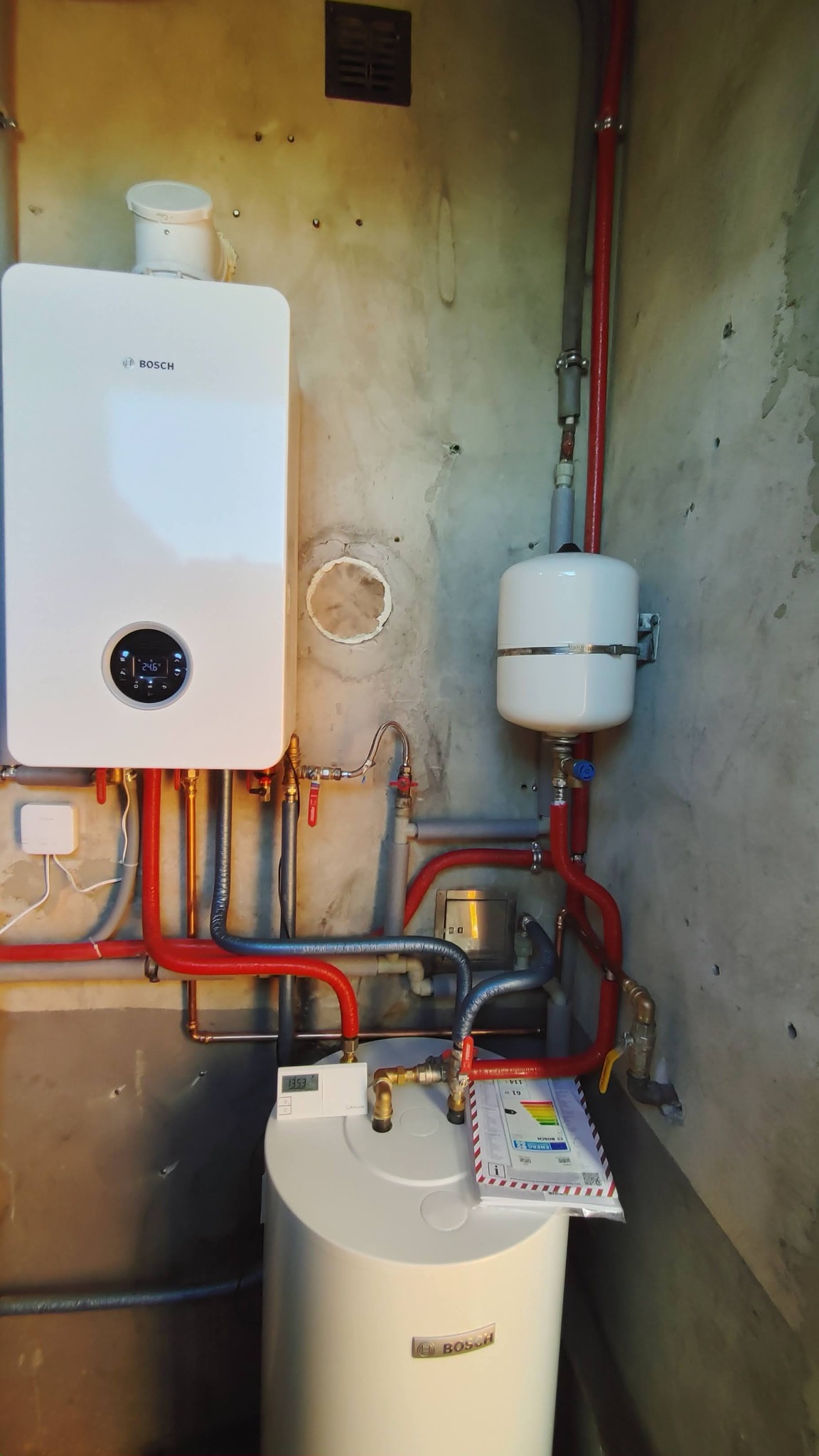 Nowoczesny, biały piec gazowy Bosch z wyświetlaczem cyfrowym i podgrzewacz wody, zamontowane na surowej ścianie z widocznymi rurami miedzianymi i plastikowymi w kolorze czerwonym i szarym,...