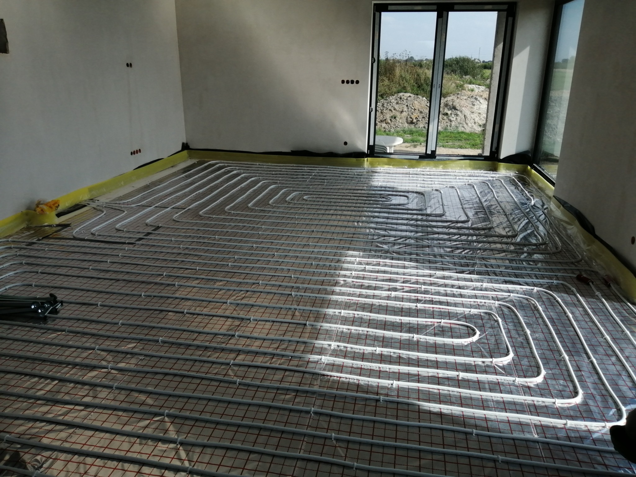 Białe rury ogrzewania podłogowego ułożone na siatce w jasnym pomieszczeniu z widokiem na zewnątrz przez okno, folia ochronna częściowo rozłożona.