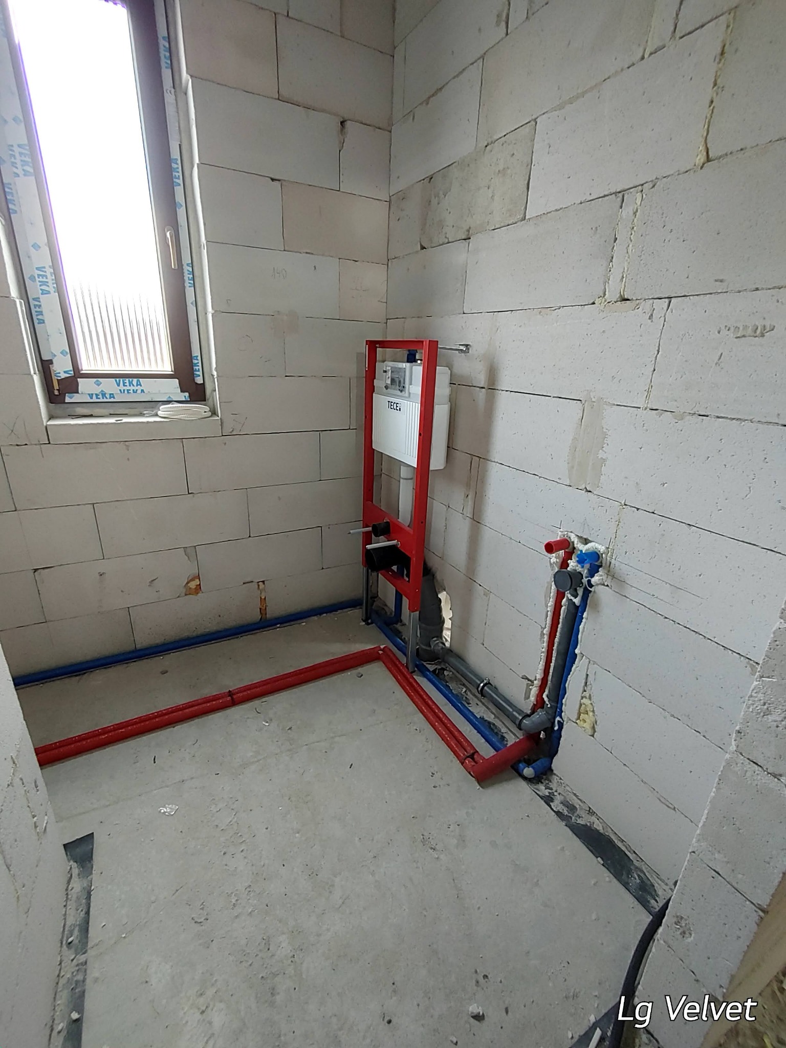 Surowy stan łazienki z widoczną instalacją podtynkową WC na czerwonym stelażu, rurami wodnymi z wyprowadzeniami na ścianie z betonu komórkowego oraz oknem z naklejkami producenta.