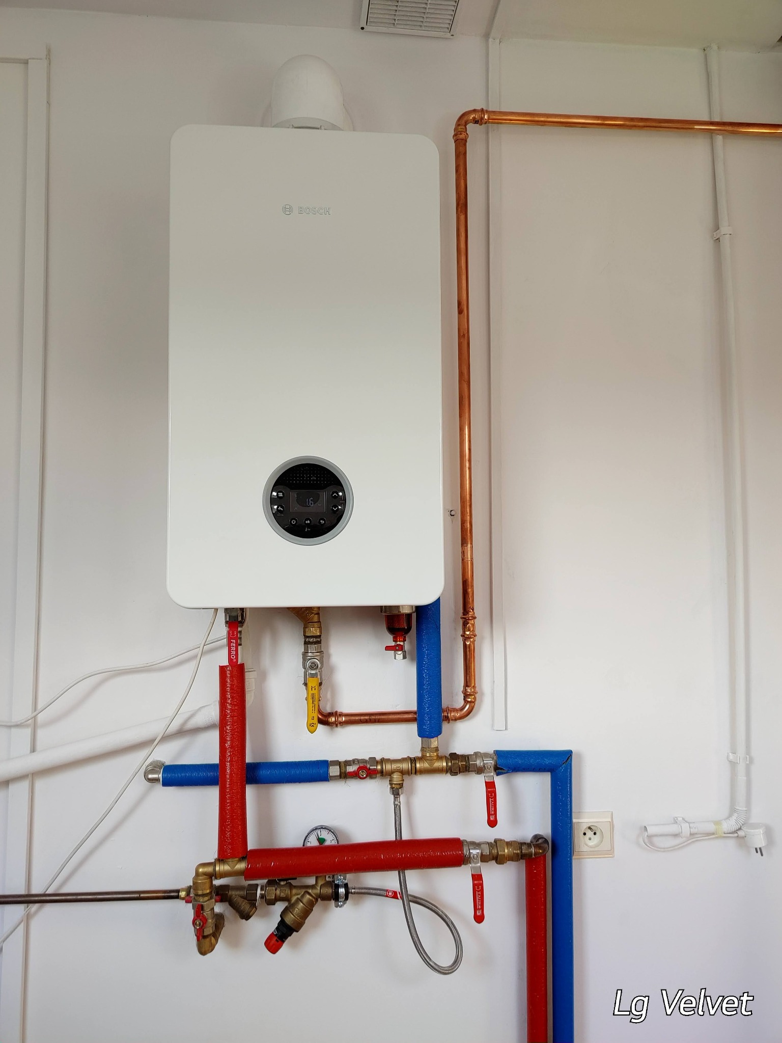Biały, wiszący kondensacyjny piec gazowy marki Bosch z miedzianymi rurami, zaworami i izolacją termiczną w kolorze czerwonym i niebieskim na białej ścianie.