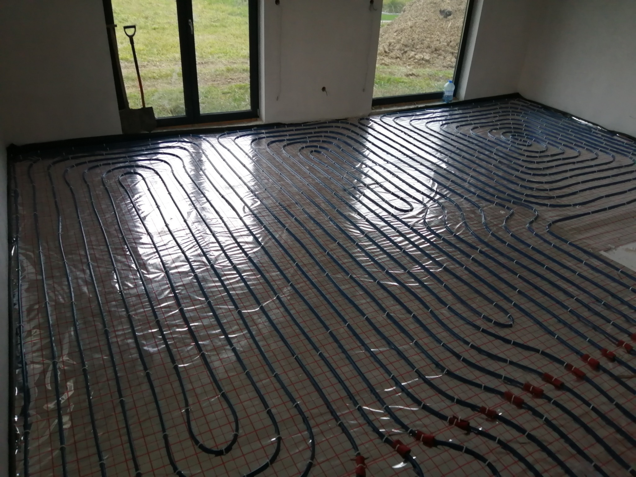 Rozłożone rury ogrzewania podłogowego na folii z siatką, widoczne złącza, w tle okno z widokiem na trawnik i łopata oparta o ścianę.