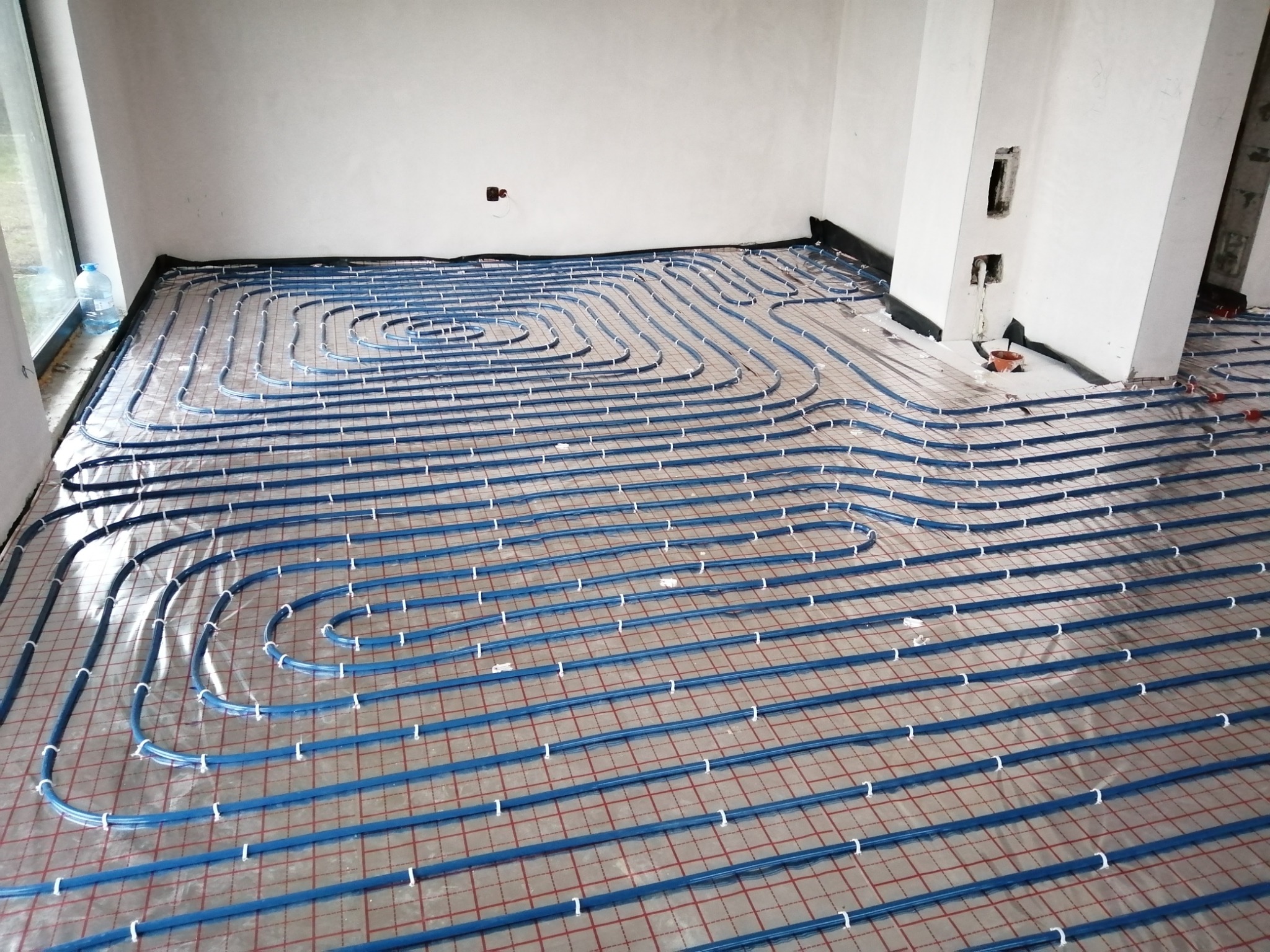Niebieskie rury ogrzewania podłogowego rozłożone spiralnie na folii z czerwoną siatką w surowym wnętrzu z białymi ścianami i niedokończonym kominem.