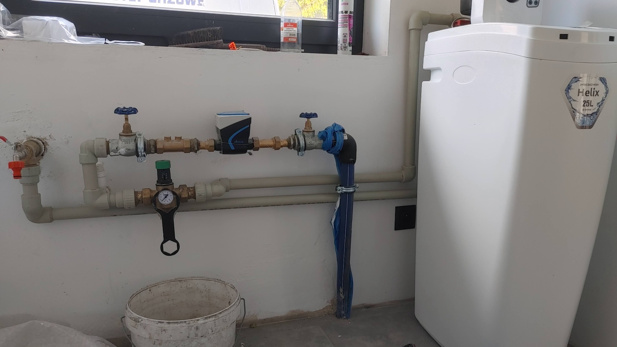 Biały zmiękczacz wody Helix 25L podłączony do instalacji hydraulicznej z zaworami, manometrem i niebieskim zabezpieczeniem rury w pomieszczeniu z oknem i wiadrem na podłodze.