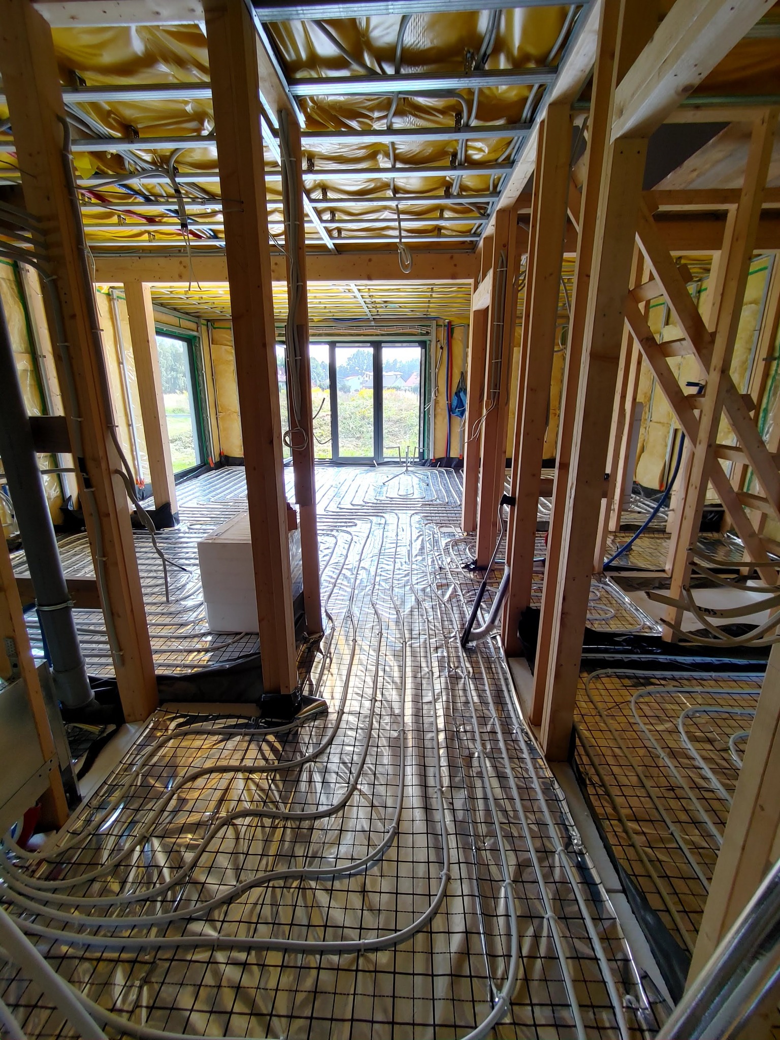 Montaż ogrzewania podłogowego w nowym domu, widoczne rury grzewcze ułożone na metalowej siatce, drewniana konstrukcja szkieletowa budynku, żółta folia izolacyjna na ścianach.