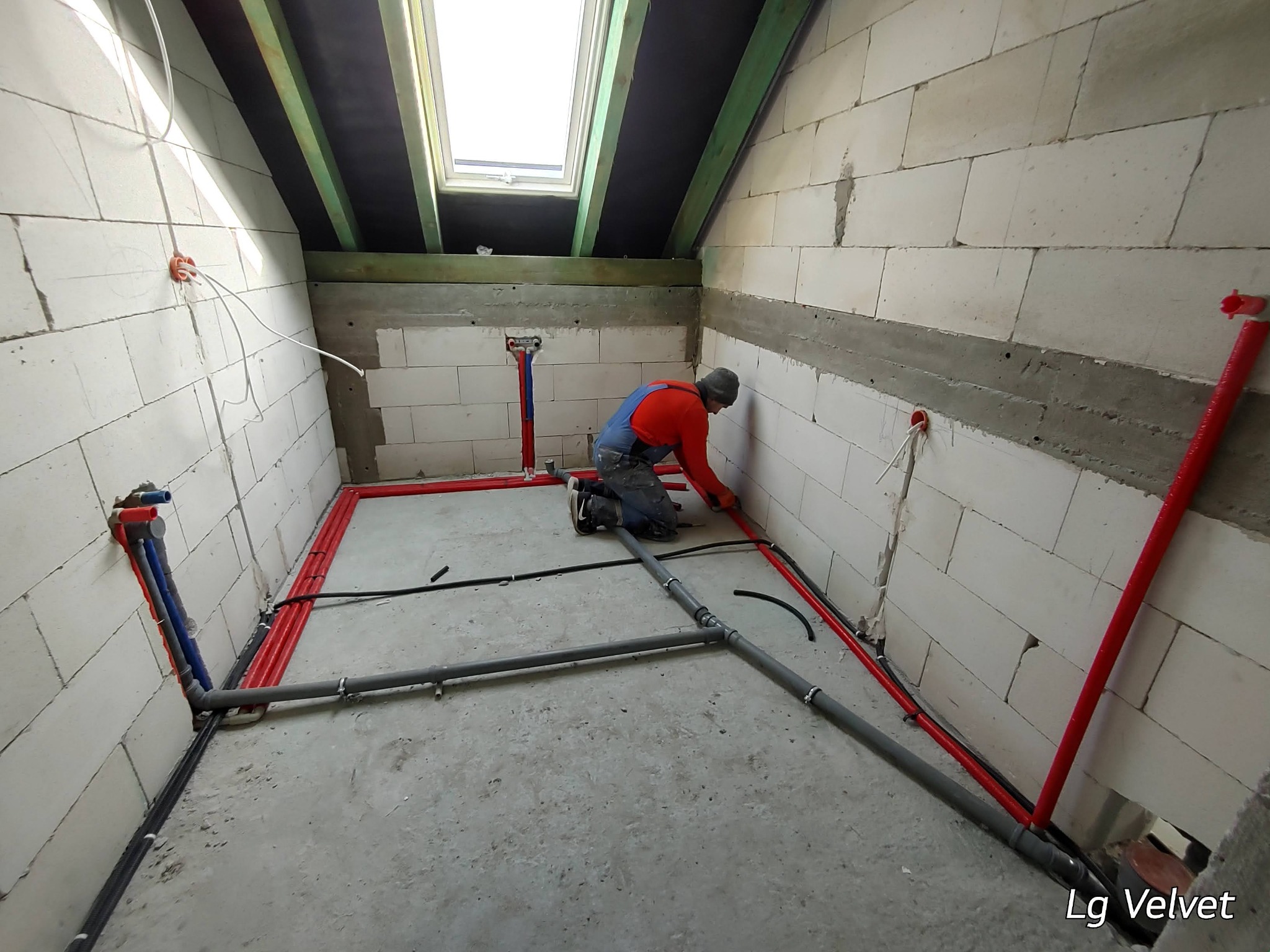 Instalator w trakcie montażu instalacji wodno-kanalizacyjnej w surowym pomieszczeniu z oknem dachowym, widoczne rury w kolorze szarym i czerwonym oraz białe ściany z betonu komórkowego.