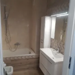 Jasna łazienka z wanną, umywalką z szafką i wysoką szafką boczną, wykończona płytkami w odcieniach beżu i brązu, z drewnopodobną podłogą.