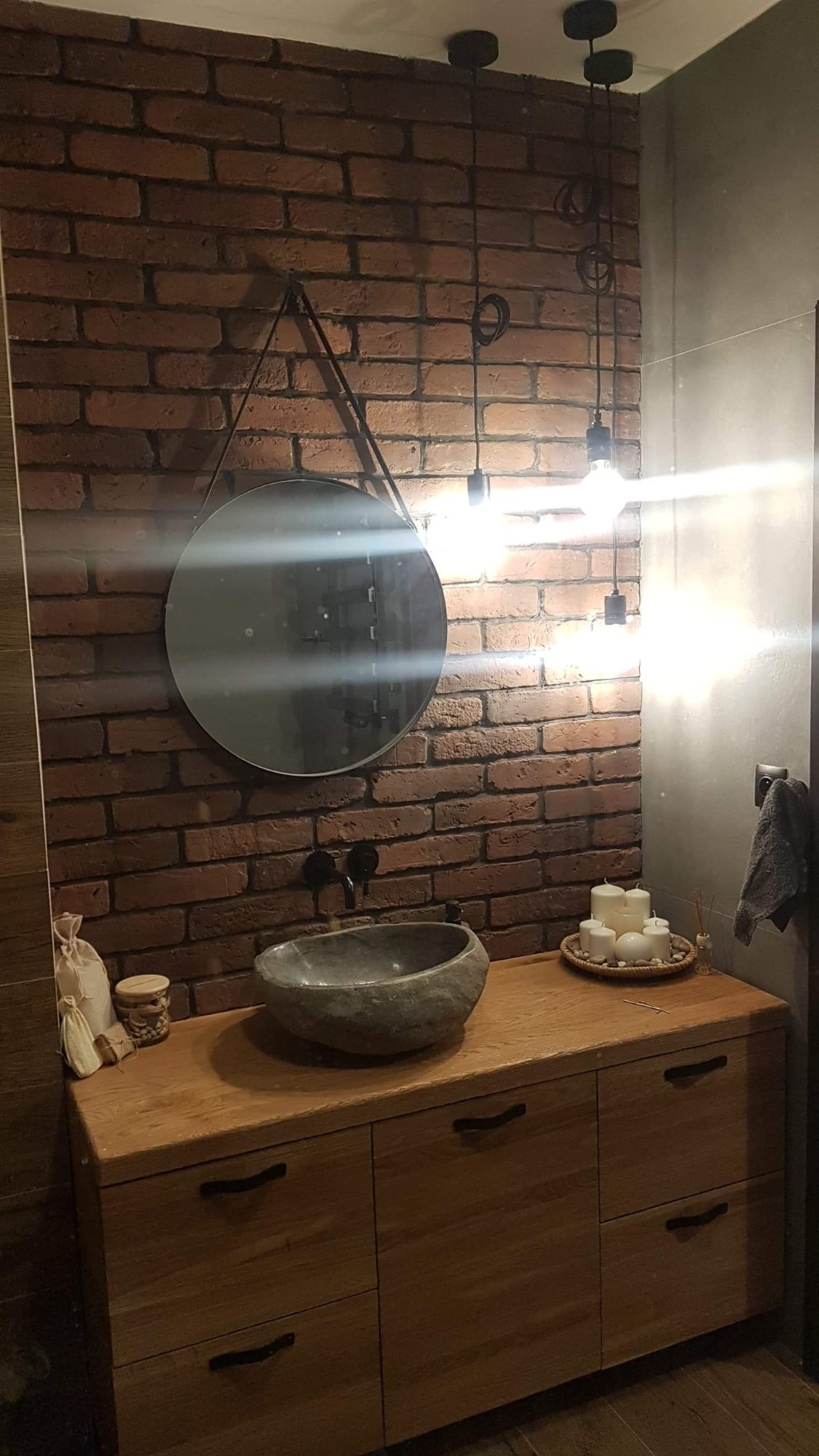 Rustykalna łazienka z umywalką w kształcie kamienia, okrągłym lustrem na skórzanym pasku i ścianą z czerwonej cegły, oświetlona wiszącymi żarówkami bez kloszy.