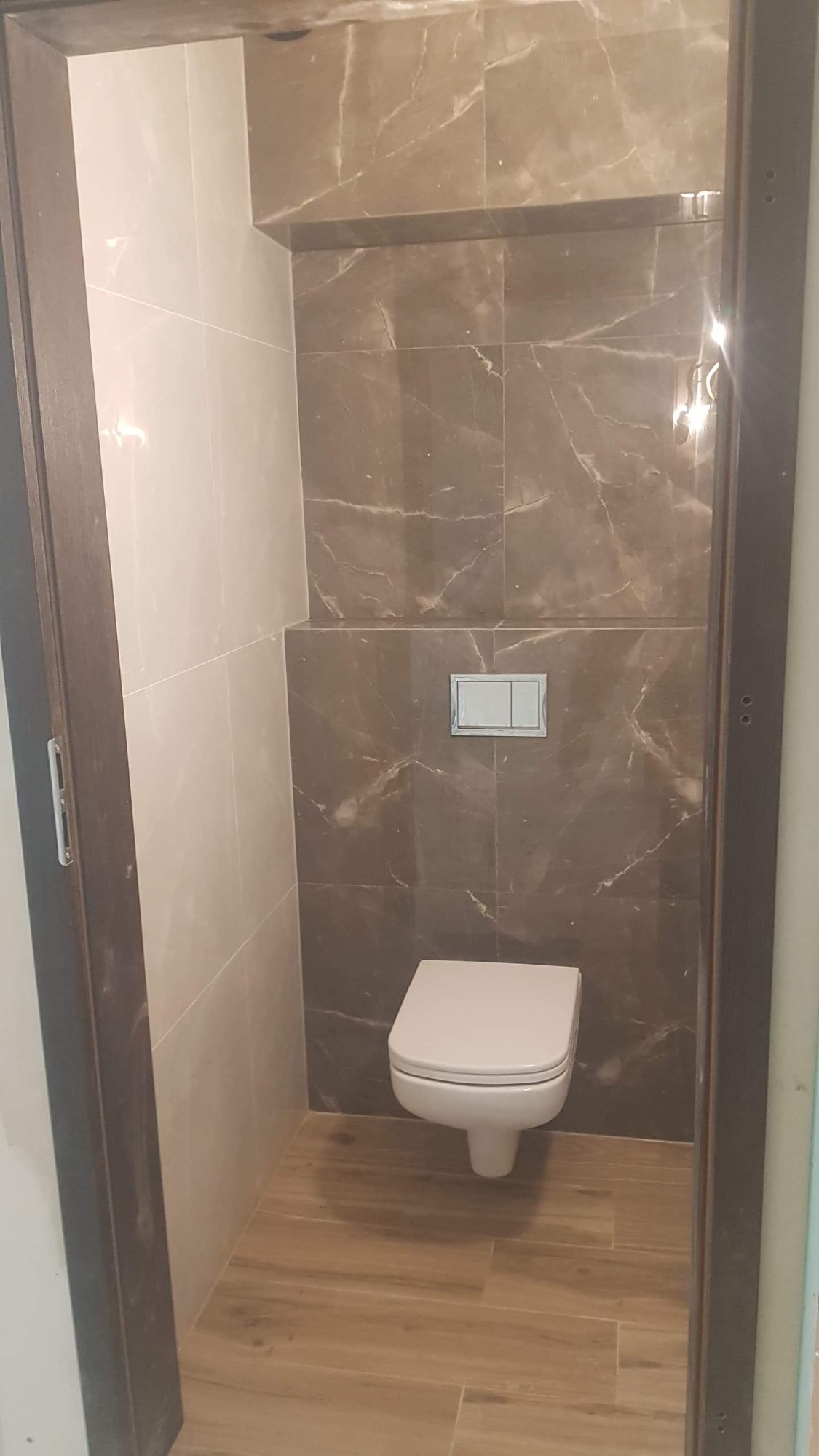 Nowoczesna toaleta podwieszana w małej łazience wykończonej płytkami imitującymi marmur w odcieniach beżu i brązu, z drewnopodobną podłogą i chromowanym przyciskiem spłukującym.