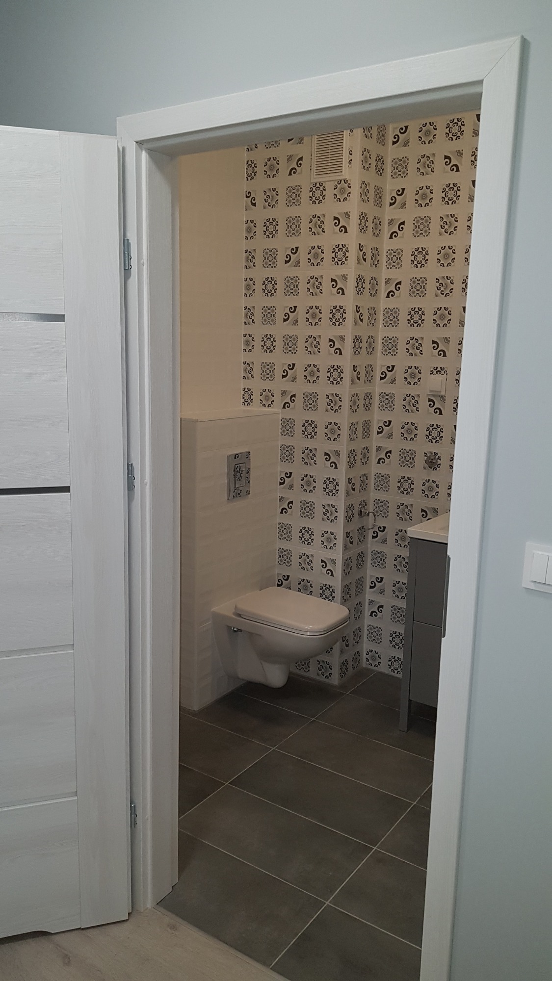 Wykończona łazienka z wiszącą toaletą, wzorzystymi płytkami na ścianie i szarą podłogą, widziana zza otwartych, białych drzwi.