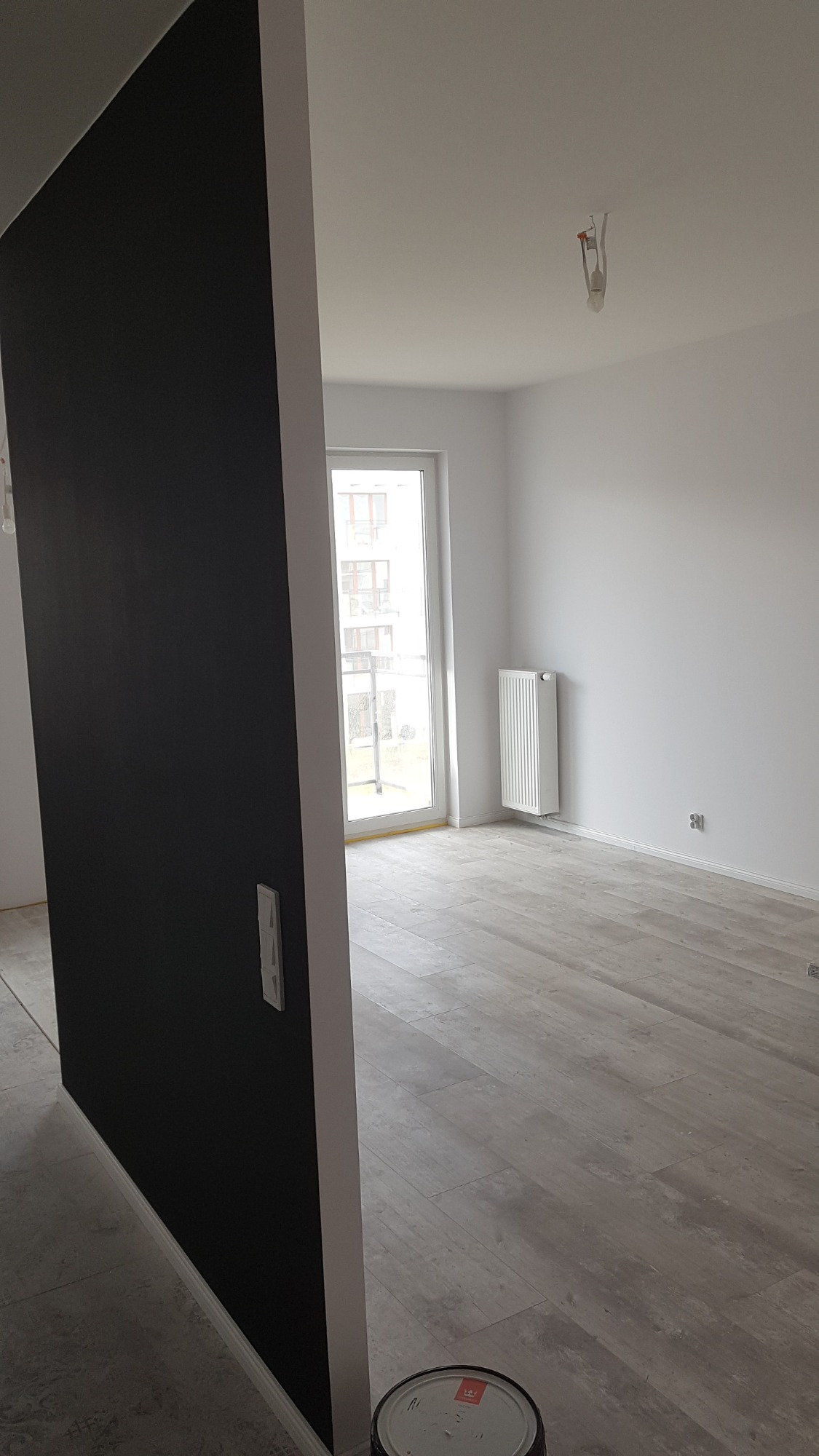 Pomalowany na czarno fragment ściany w nowym mieszkaniu, widok na jasne pomieszczenie z grzejnikiem i oknem balkonowym, na podłodze panele imitujące drewno, w rogu puszka farby.
