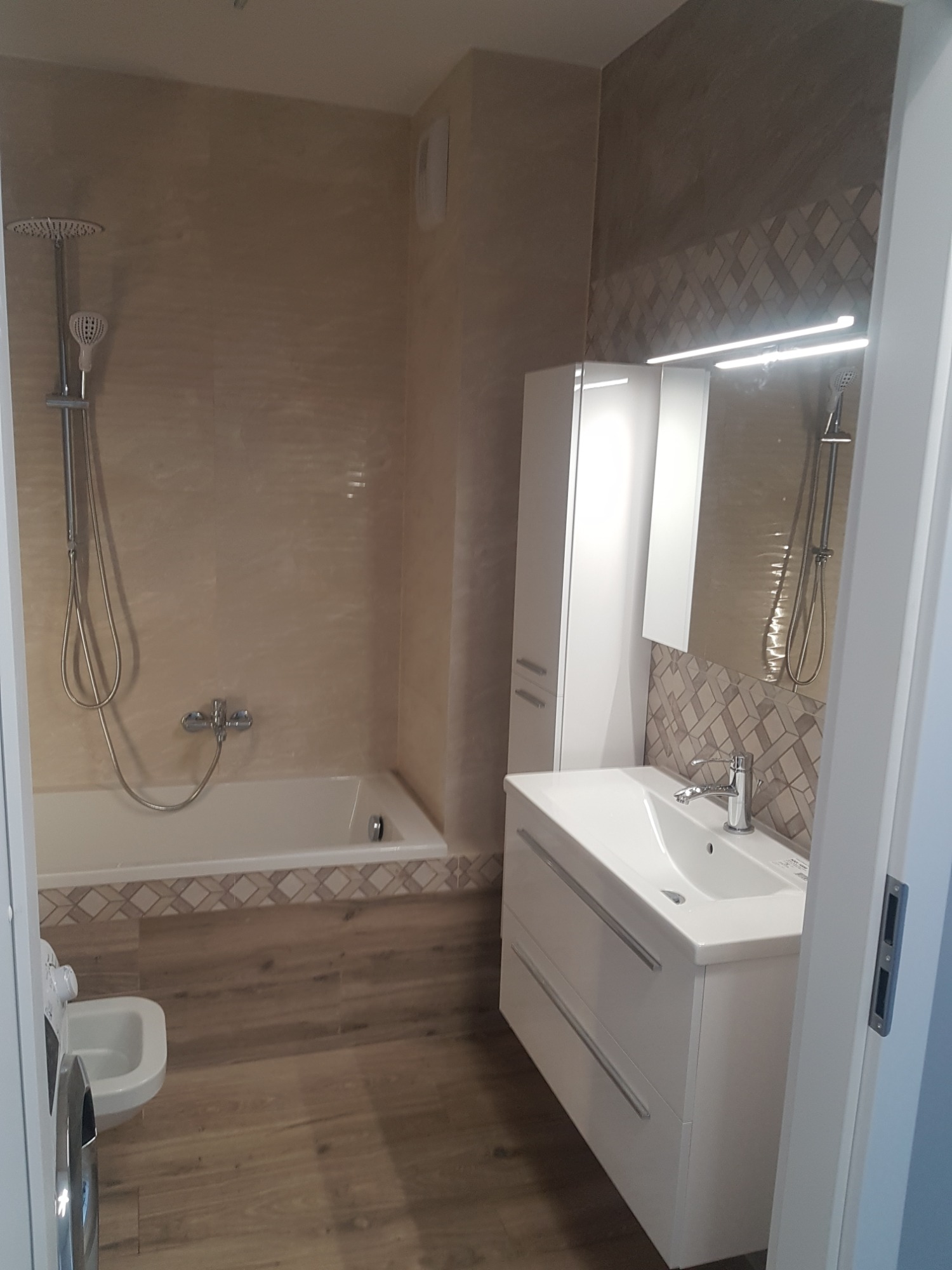 Jasna łazienka z wanną, umywalką z szafką i wysoką szafką boczną, wykończona płytkami w odcieniach beżu i brązu, z drewnopodobną podłogą.