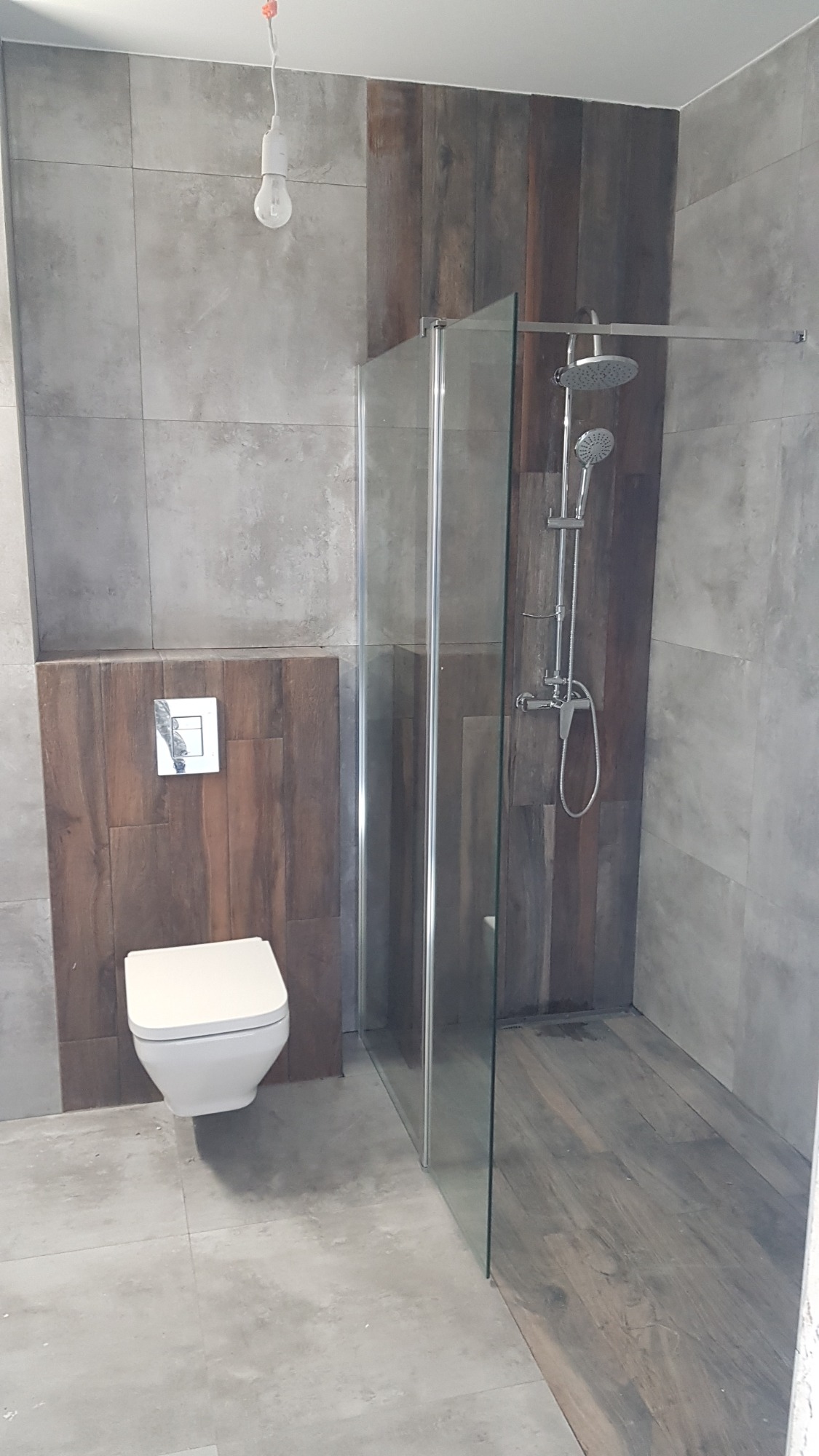 Nowoczesna łazienka z kabiną prysznicową typu walk-in, toaletą podwieszaną i okładziną ścienną imitującą drewno, surowa żarówka zwisająca z sufitu.