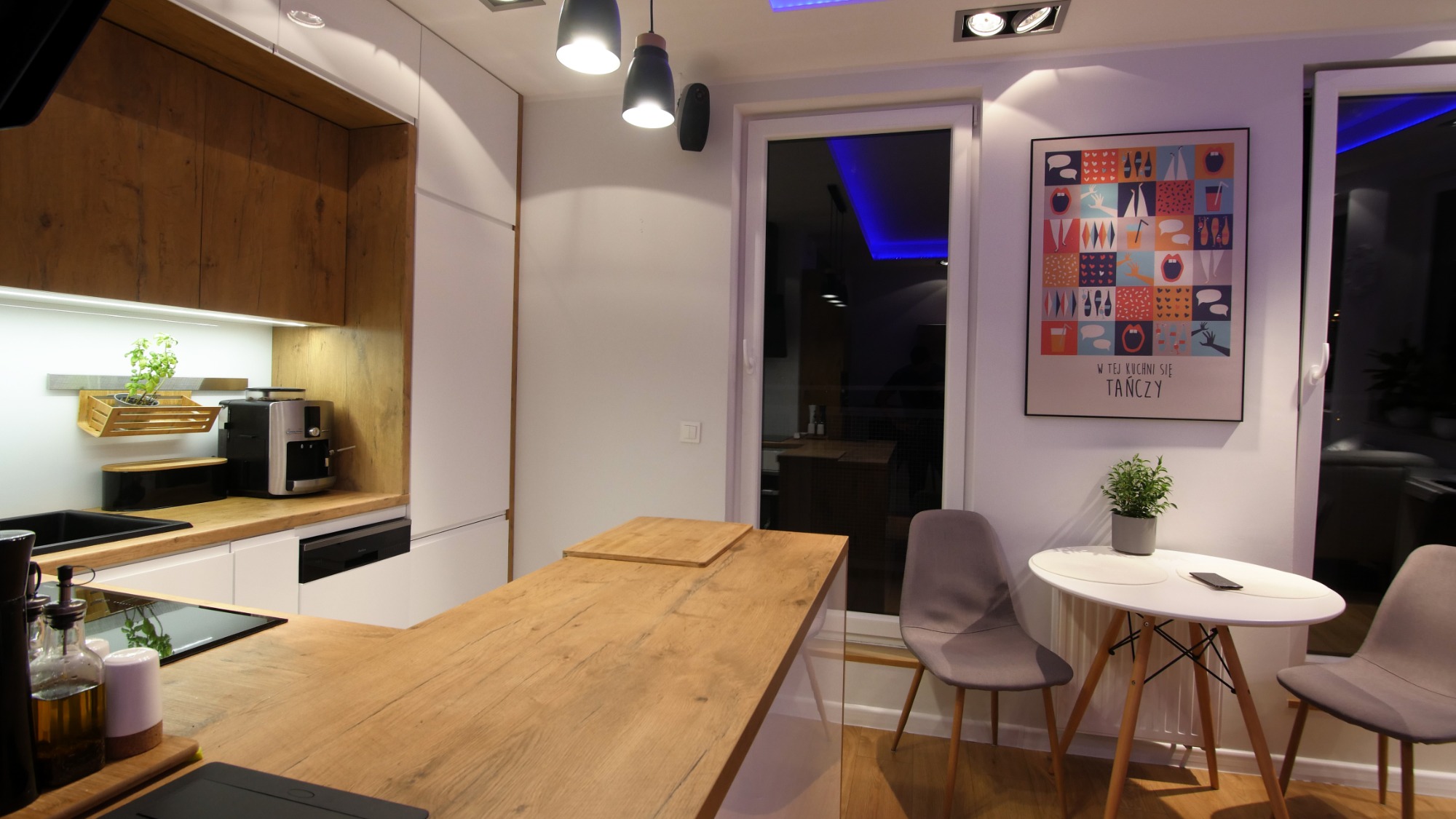 Nowoczesna kuchnia z drewnianym blatem, białymi szafkami i oświetleniem LED. Na blacie drewniana deska do krojenia i czarny zlew. Na ścianie plakat z napisem 'W tej kuchni się tańczy'.
