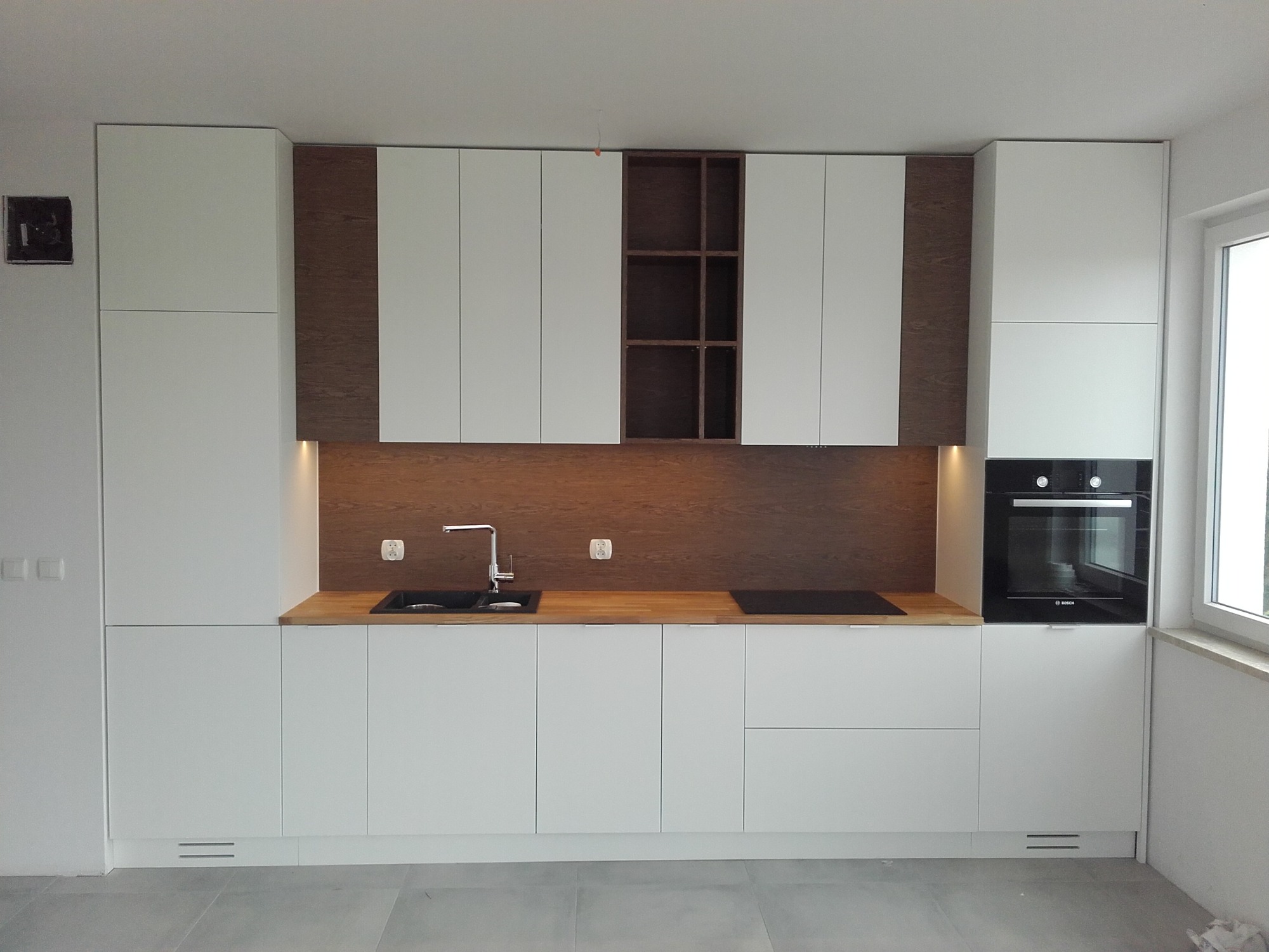Nowoczesna, minimalistyczna kuchnia z białymi szafkami, drewnianym blatem i brązowym panelem ściennym, czarny zlewozmywak i płyta indukcyjna, zabudowany piekarnik marki Bosch po prawej stronie okna.