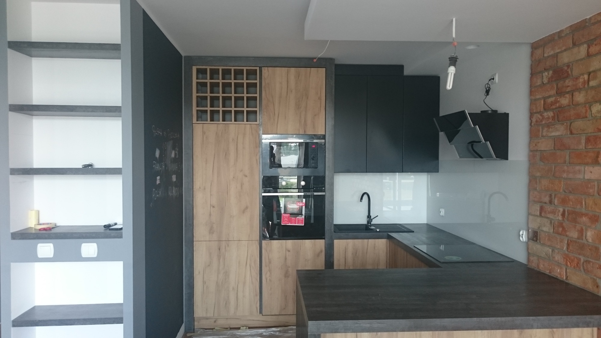 Nowoczesna kuchnia z drewnianymi frontami szafek, czarnymi elementami i ceglaną ścianą, widoczna tablica kredowa z napisem i otwarte półki.