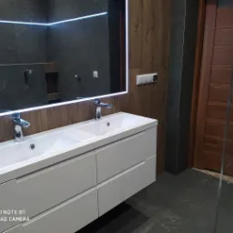 Nowoczesna łazienka z podwójną białą umywalką nablatową, lustrem z podświetleniem LED, ciemnymi płytkami imitującymi kamień i drewnianymi akcentami na ścianie oraz drzwiami.