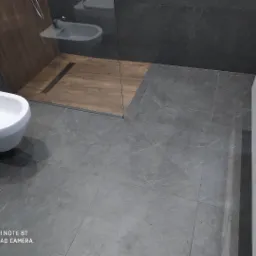 Nowoczesna łazienka z szarymi płytkami imitującymi kamień, białą bidetą i szafką, oraz kabiną prysznicową z drewnopodobną podłogą i odpływem liniowym.