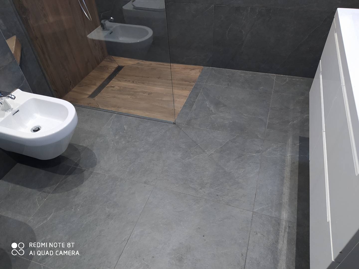 Nowoczesna łazienka z szarymi płytkami imitującymi kamień, białą bidetą i szafką, oraz kabiną prysznicową z drewnopodobną podłogą i odpływem liniowym.