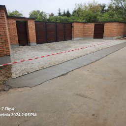 Craftmator Sp. z o.o. - Fragment podjazdu z kostki granitowej, częściowo zalany świeżym betonem, zabezpieczony taśmą ostrzegawczą, otoczony ceglanym murem z drewnianą bramą w tle.