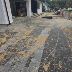 Craftmator Sp. z o.o. - Częściowo ułożona, szara kostka brukowa przed domem jednorodzinnym, piasek rozsypany na powierzchni, widoczne narzędzia i pracownicy w tle.