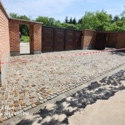 Craftmator Sp. z o.o. - Ułożona kostka granitowa na podjeździe z bramą wjazdową z czerwonej cegły, zabezpieczona czerwoną taśmą ostrzegawczą, widoczne resztki zaprawy i asfaltu.