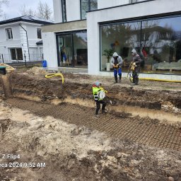 Craftmator Sp. z o.o. - Przygotowanie zbrojenia w wykopie pod fundamenty domu jednorodzinnego; pracownicy w odzieży roboczej i kaskach, widoczny fragment budynku z dużymi oknami.