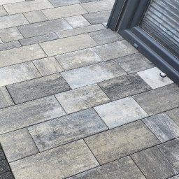 Craftmator Sp. z o.o. - Ułożona szara kostka brukowa o prostokątnym kształcie przylegająca do ciemnej ramy okiennej i obrzeża z kostki brukowej, widoczne źdźbła trawy.