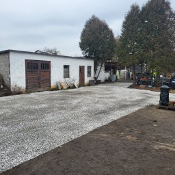 Craftmator Sp. z o.o. - Podwórko z budynkami gospodarczymi i nowo utwardzoną drogą żwirową, widoczne drzewa, samochód osobowy i elementy budowlane zabezpieczone folią.