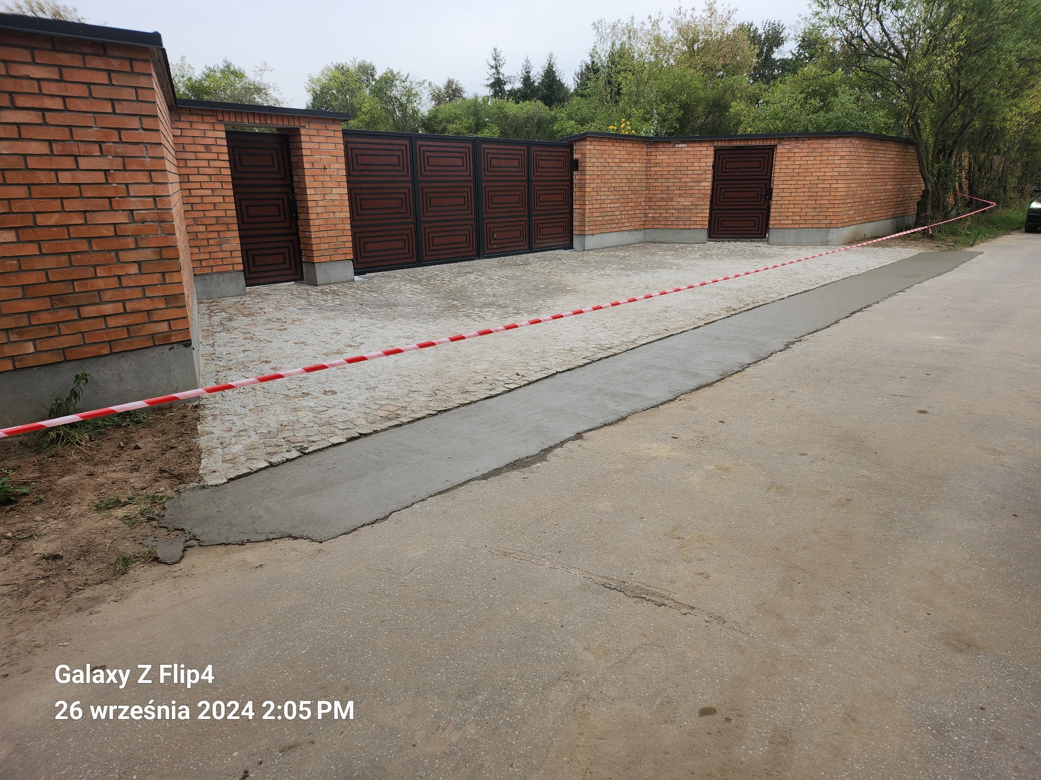 Fragment podjazdu z kostki granitowej, częściowo zalany świeżym betonem, zabezpieczony taśmą ostrzegawczą, otoczony ceglanym murem z drewnianą bramą w tle.