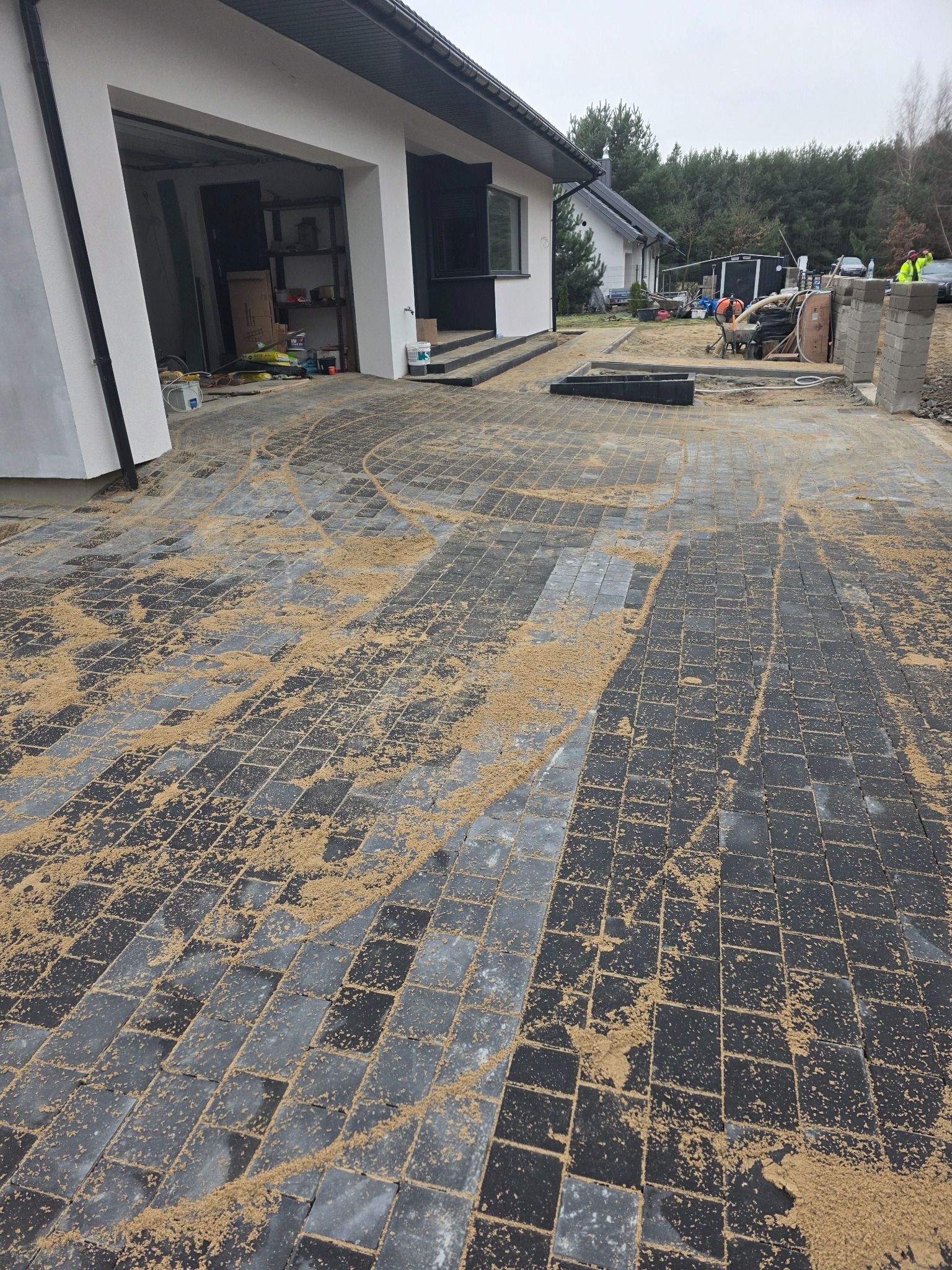 Częściowo ułożona, szara kostka brukowa przed domem jednorodzinnym, piasek rozsypany na powierzchni, widoczne narzędzia i pracownicy w tle.