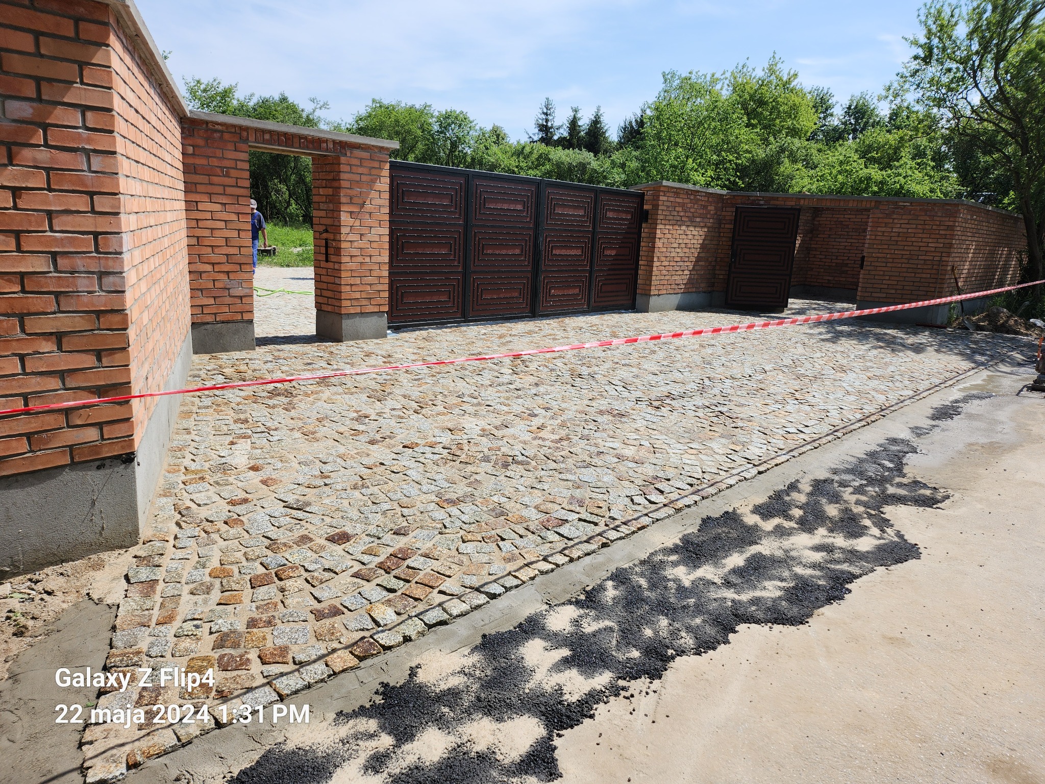 Ułożona kostka granitowa na podjeździe z bramą wjazdową z czerwonej cegły, zabezpieczona czerwoną taśmą ostrzegawczą, widoczne resztki zaprawy i asfaltu.